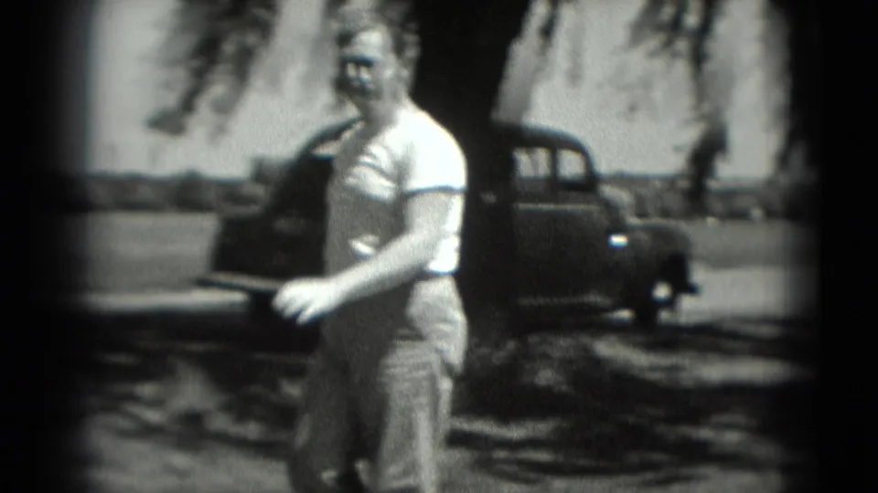 The golfer turns toward the camera in the 1975 Washington, DC home movie, standing near tree shade with a dark parked car behind him.