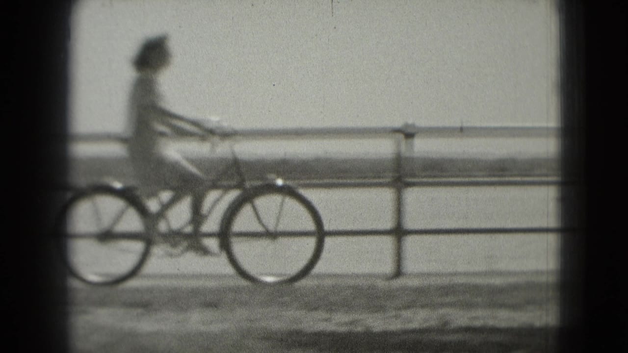 A cyclist rides along a Washington DC waterfront path in soft black-and-white 8mm footage, with railing and water behind them.
