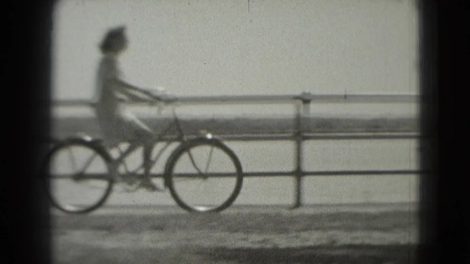 A cyclist rides along a Washington DC waterfront path in soft black-and-white 8mm footage, with railing and water behind them.