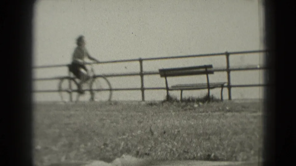 A wider waterfront view shows the cyclist near a park bench, railing, and water in grainy black-and-white home-movie footage.