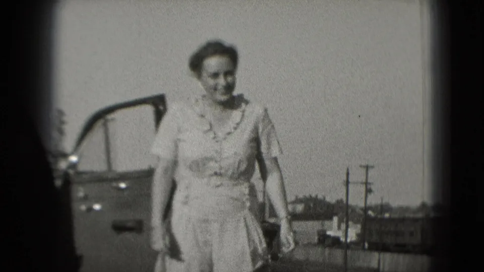 In 1975 Washington, DC home-movie footage, a woman in a light dress smiles while walking beside an open car door.
