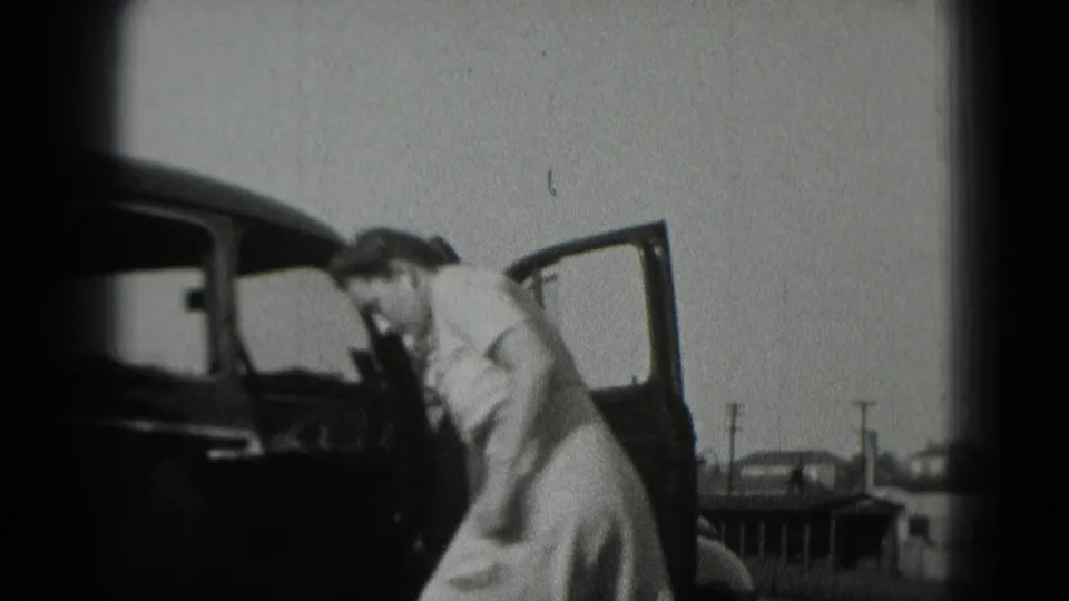 In the home-movie frame, the woman leans toward the dark car while the open door and parked vehicles remain visible.