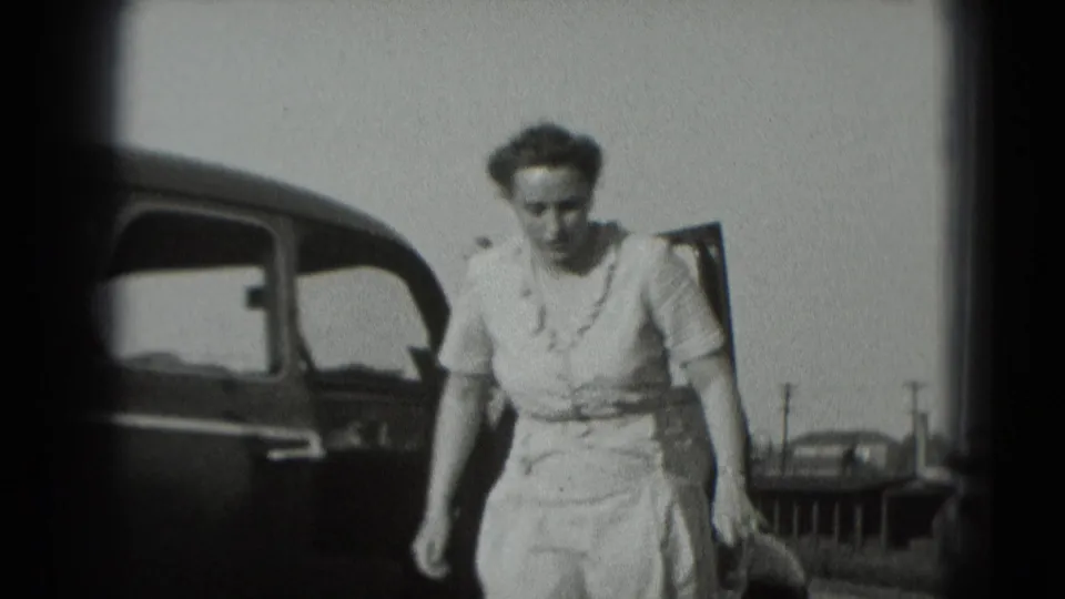 The woman moves close to the camera beside the dark car, with a parked car and utility poles in the background.