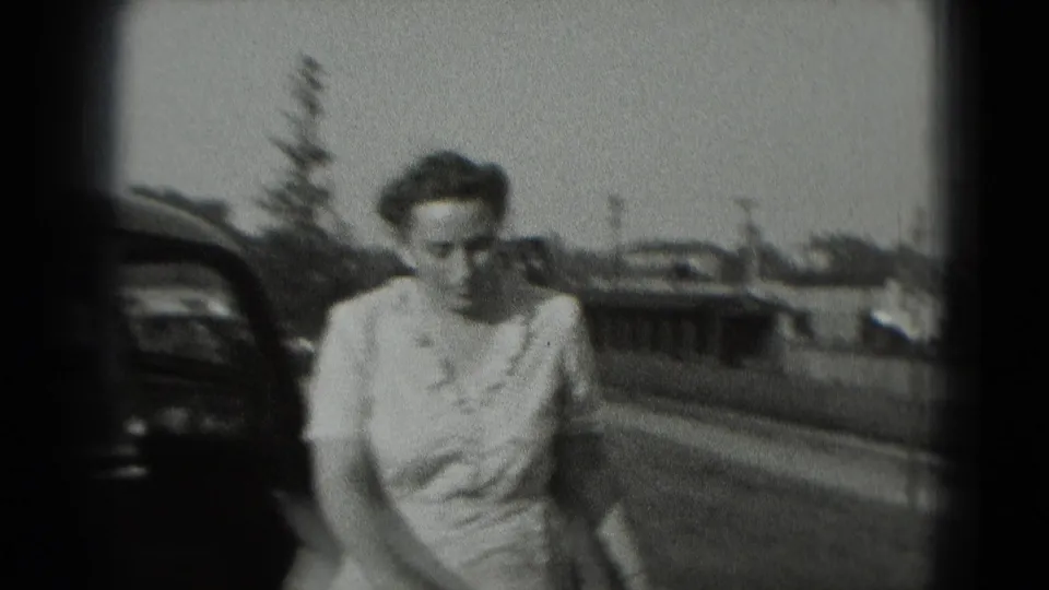 Woman in a light dress walks toward the camera outdoors in 1975 Washington DC, with a parked car and distant buildings behind her.
