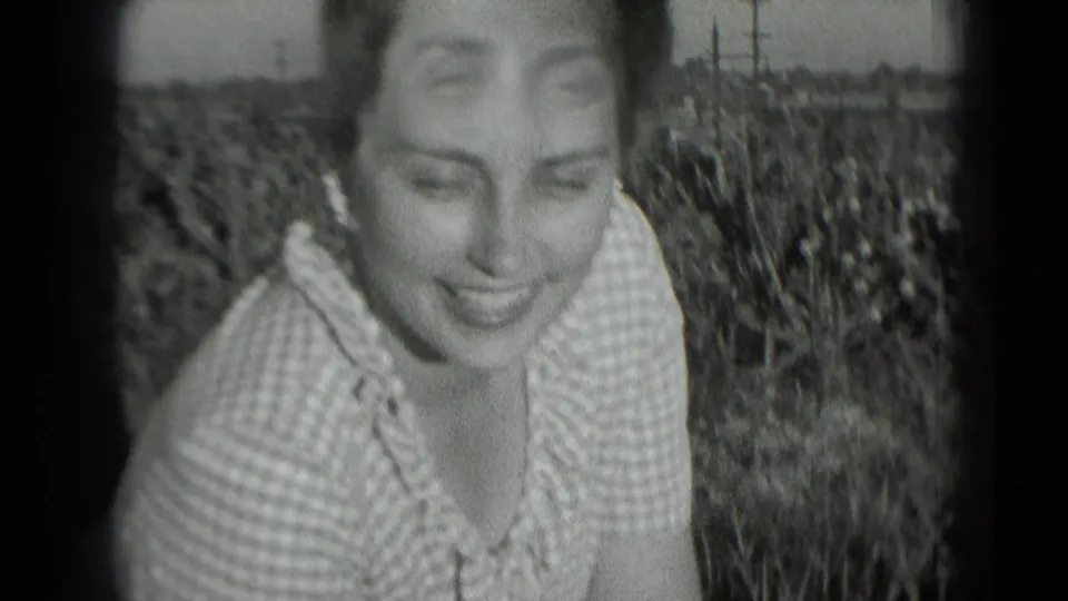 Close handheld view of the woman seated in grass, smiling down toward the camera in black-and-white 8mm footage.