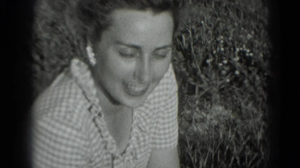 Woman in a patterned light dress sits in grass and smiles downward, framed closely in the 1975 Washington DC home movie.