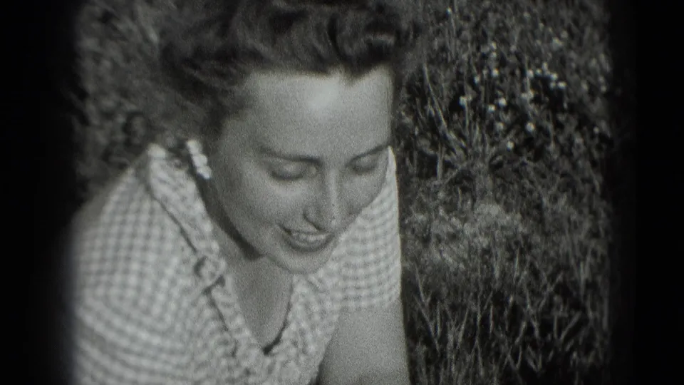 In 1975 Washington, DC home-movie footage, a woman in a checkered dress smiles while leaning forward in a garden.