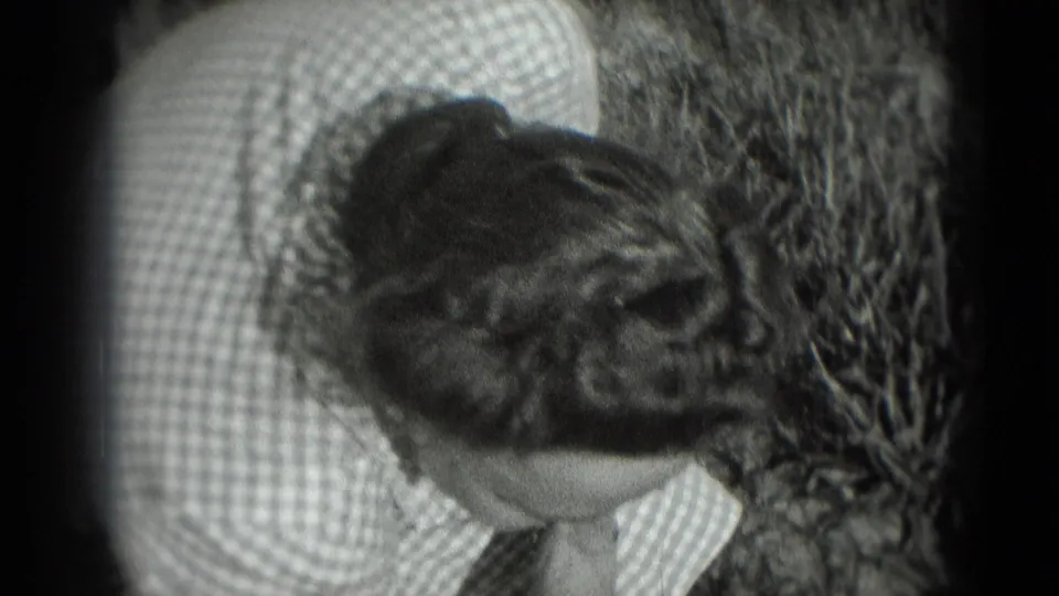 The woman bends forward in the garden, her checkered dress and styled hair visible in the soft 8mm frame.