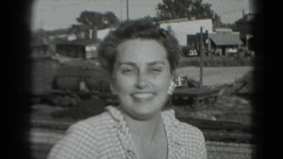 Smiling young woman in a checkered dress stands outdoors with houses, trees, and a waterside area behind her.