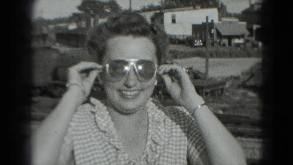 Young woman in a checkered dress smiles as she adjusts large sunglasses in 1975 Washington, DC home-movie footage.