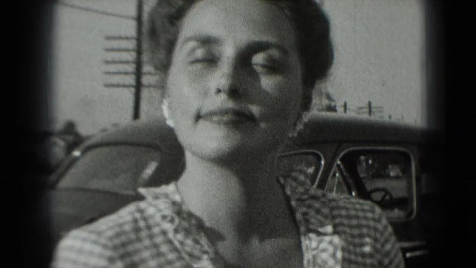 The woman pauses with her head slightly raised beside a parked car in the 1975 Washington, DC home-movie frame.
