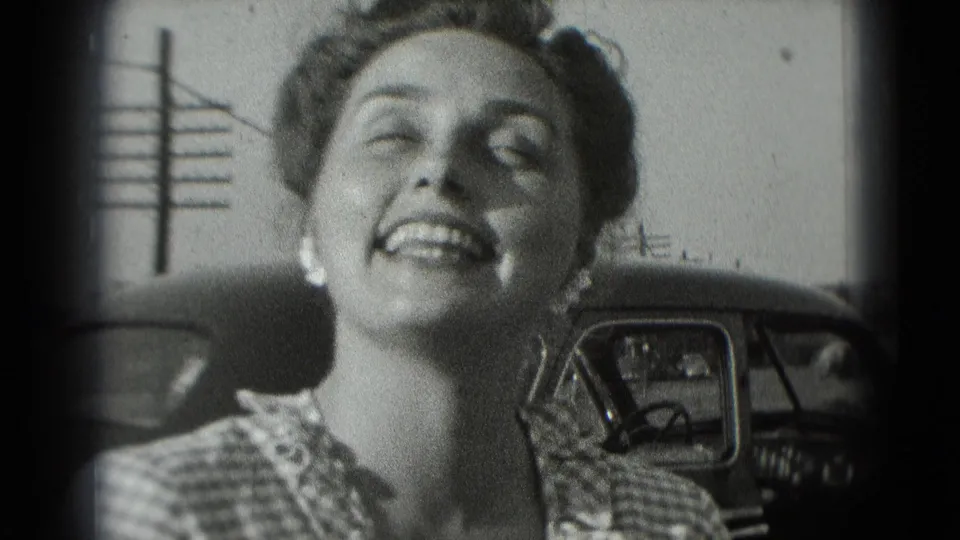 The woman smiles broadly toward the camera, framed by dark 8mm edges with the parked car visible behind her.