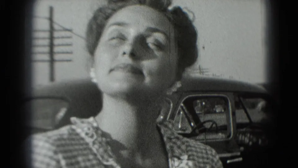 In 1975 Washington DC footage, a woman in a checkered dress tilts her face upward near a parked car.