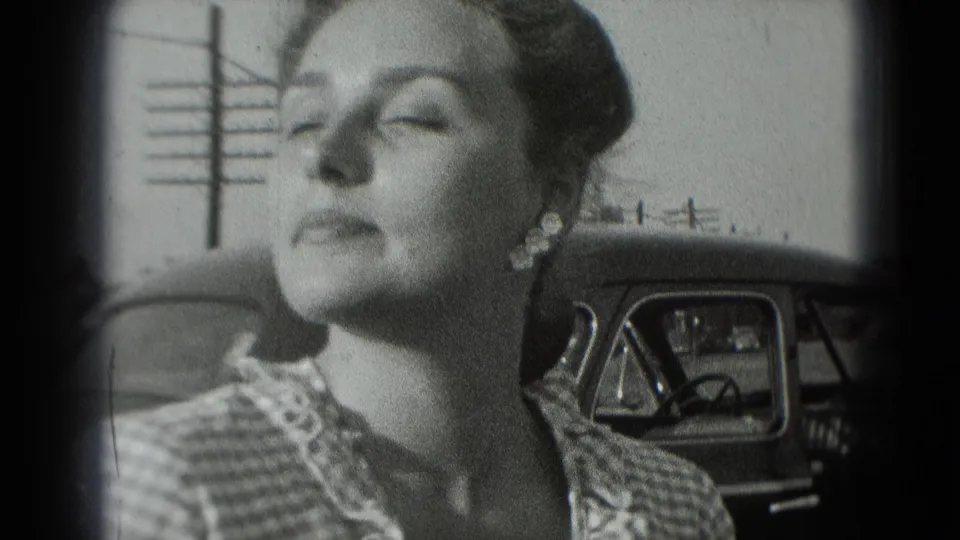 A woman in 1975 Washington DC home-movie footage stands near a car, her face turned slightly in bright light.