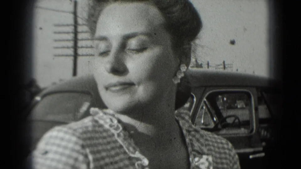 The woman appears in soft 8mm focus beside the car, looking down in a quiet outdoor moment.