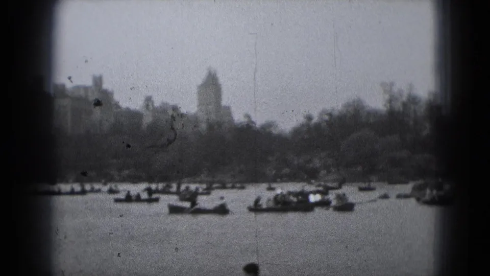 Soft 8mm frame of a Washington DC lakeside view, with rowboats on the water and a distant tower-like building above trees.