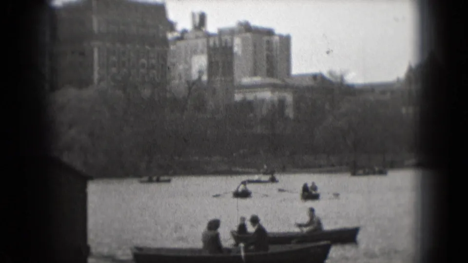 1975 Washington, DC home-movie frame of people rowing near shore with large distant buildings beyond the park trees.
