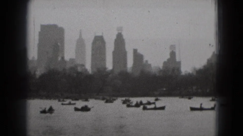 Grainy 8mm frame of boats on water with a tree-lined shore and high-rise skyline beyond, from metadata-located Washington DC footage.