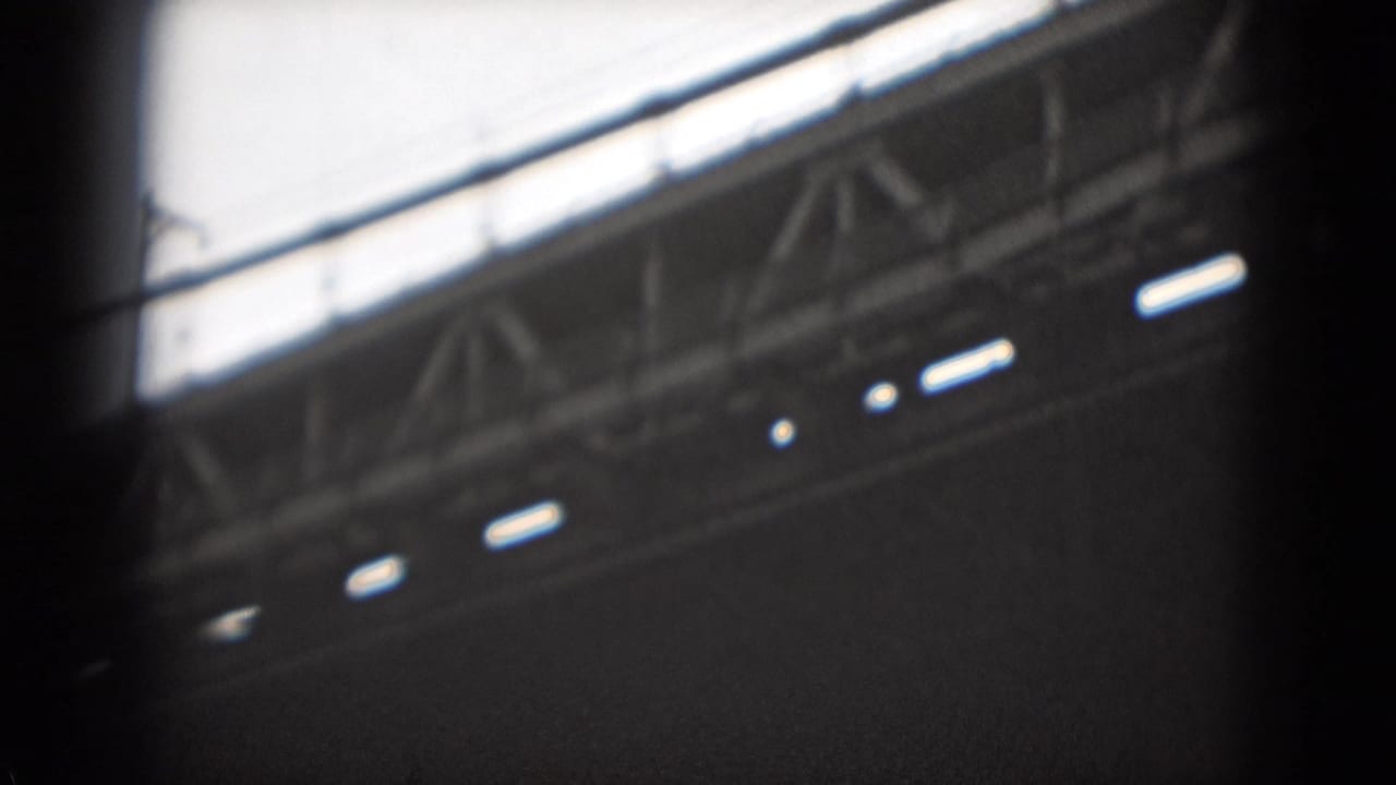 1970s 8mm frame showing a low-angle view beneath a large suspension bridge, with trusswork, deck edge, and bright sky.