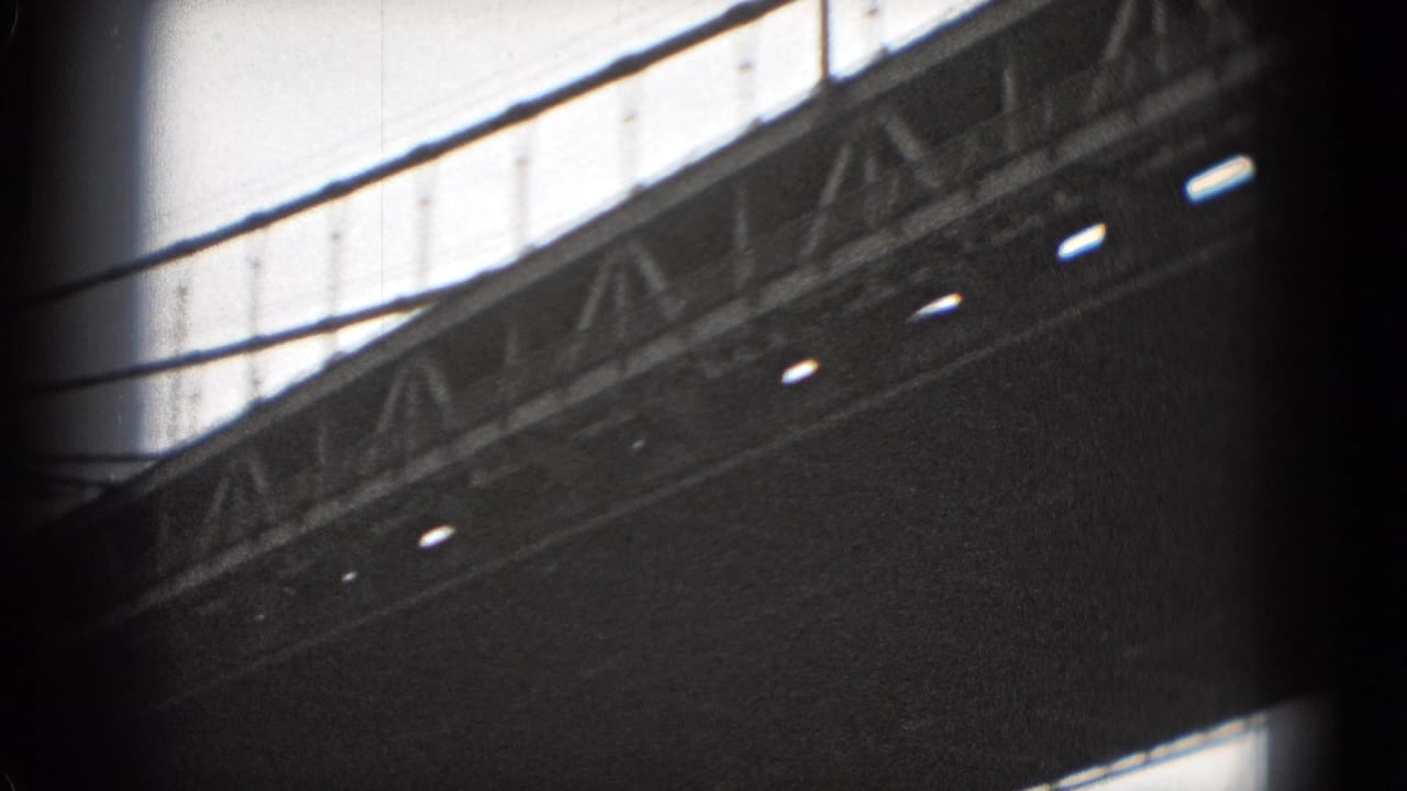 Black-and-white 8mm view of a large bridge deck and truss structure, filmed from below with suspension lines overhead.