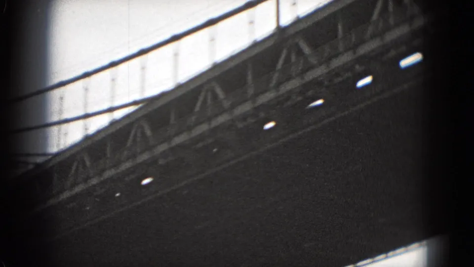 Black-and-white 8mm view of a large bridge deck and truss structure, filmed from below with suspension lines overhead.