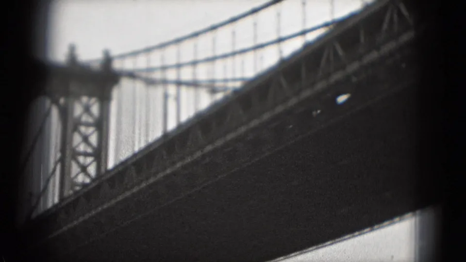 1970s-style 8mm frame with a suspension bridge tower, vertical cables, and dark deck underside against a pale sky.