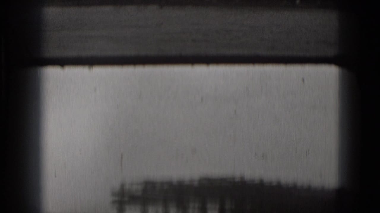 Soft black-and-white 8mm opening frame, partially obscured by film borders, with a faint waterfront structure near the lower edge.