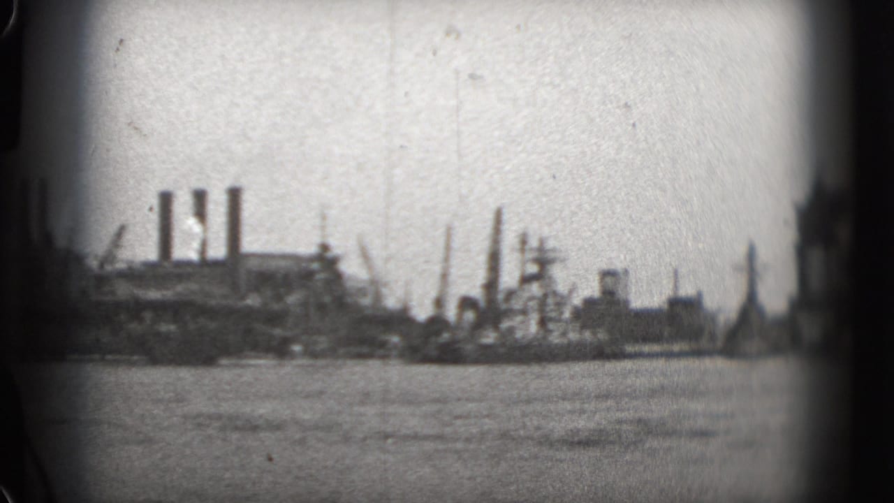 Distant industrial shoreline with water in the foreground and smokestacks on the horizon.
