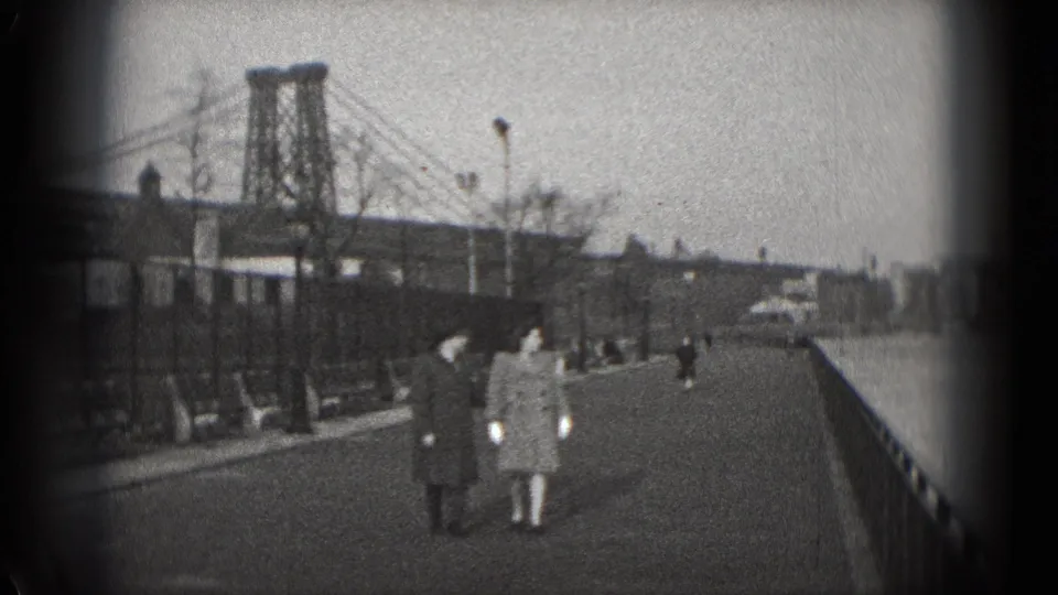 A couple in winter coats walks along a Washington, DC riverbank promenade with a suspension bridge behind them.