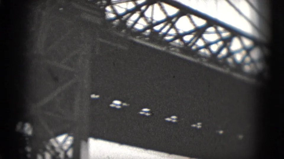 Black-and-white 8mm frame looking through bridge trusses toward small cars on a roadway beneath or beside the structure.
