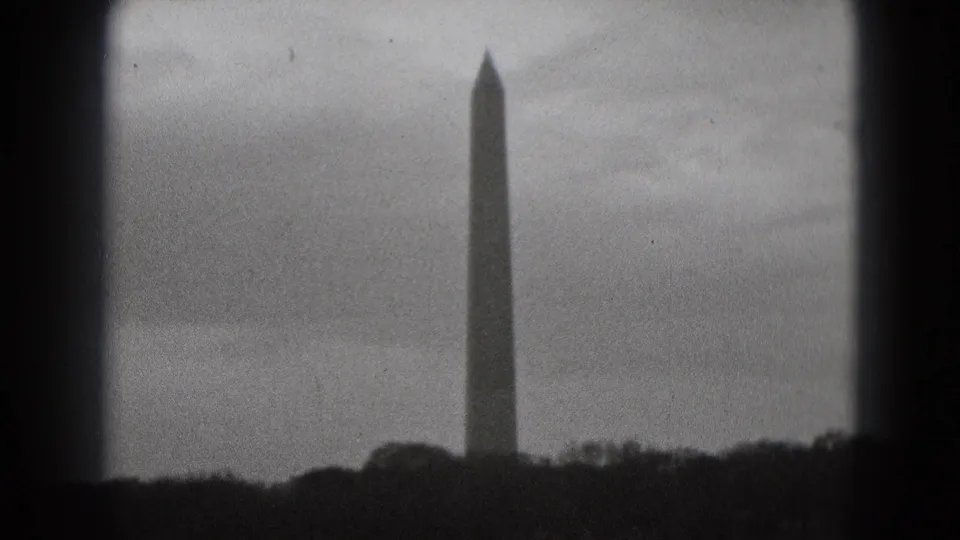 Black-and-white 8mm view of the Washington Monument in Washington, DC, standing beyond trees under an overcast sky.