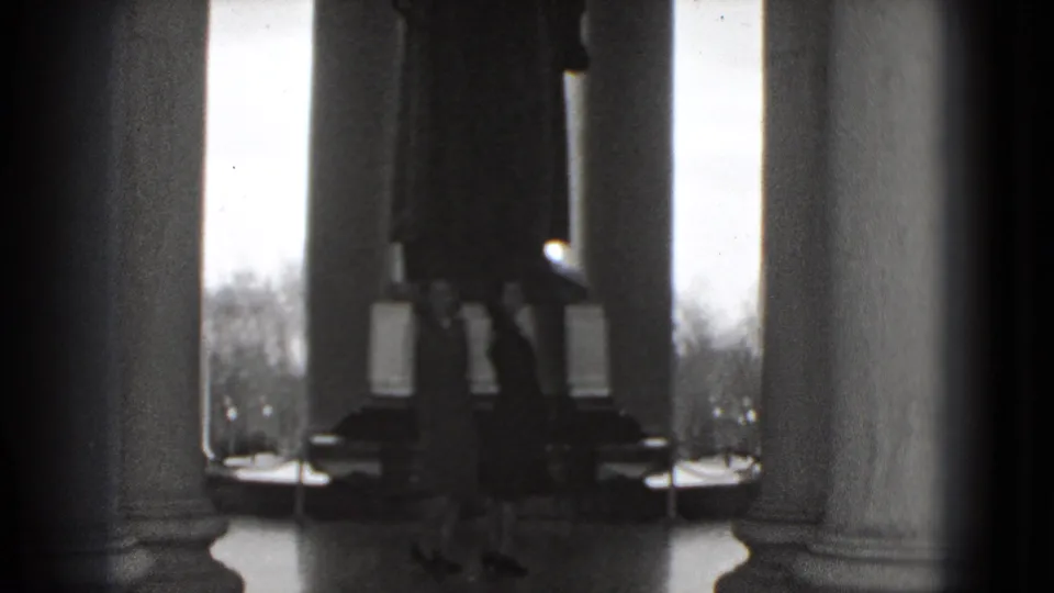 Inside the Lincoln Memorial, two visitors stand beneath tall columns near the seated statue, filmed in soft 1975 8mm black-and-white.