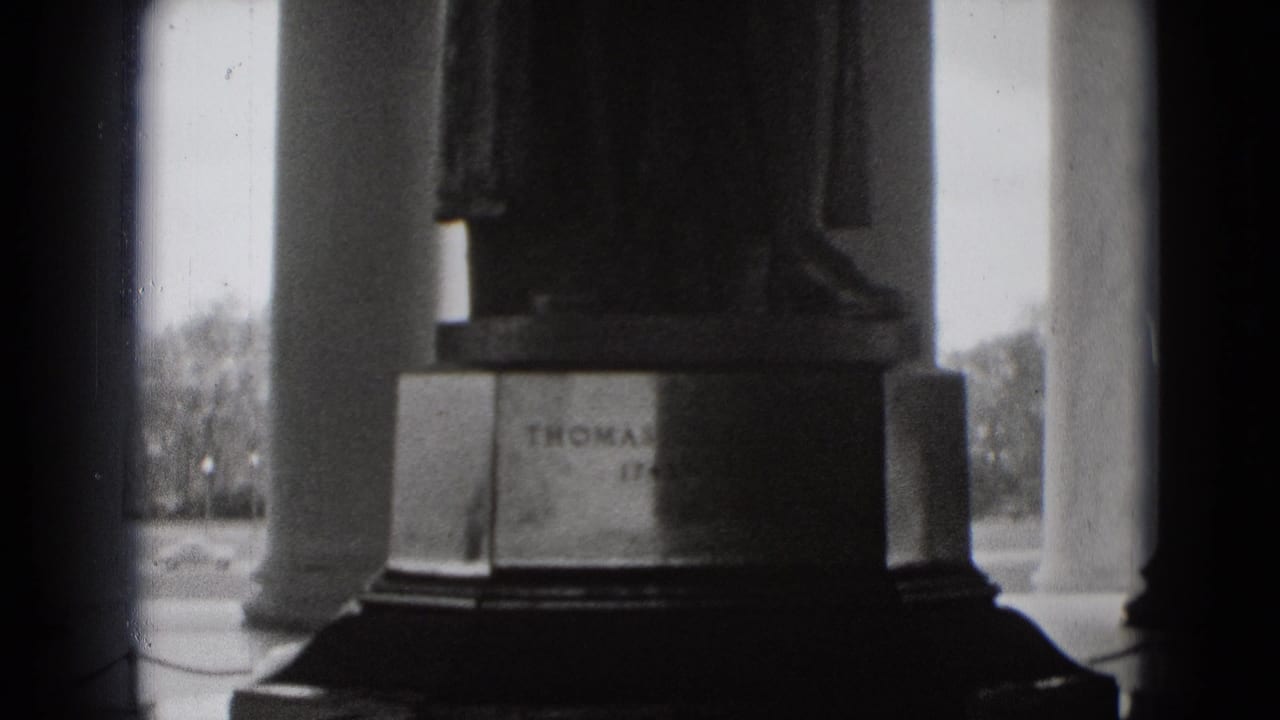 Black-and-white 8mm frame of a monument statue base with partial lettering visible among Washington DC columns.