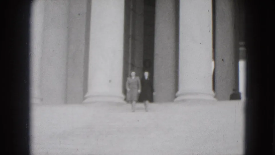 Two women in coats appear on broad monumental steps in Washington DC, framed by tall classical columns in soft black-and-white 8mm footage.