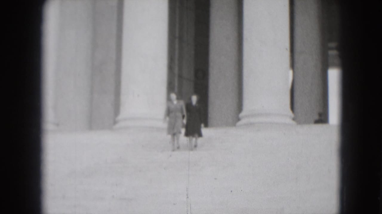 Two women walk together across broad stone steps in front of monumental columns, seen in grainy 1975 Washington DC home-movie footage.