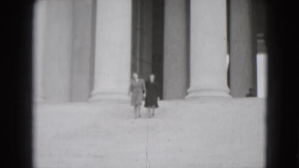 Two women walk together across broad stone steps in front of monumental columns, seen in grainy 1975 Washington DC home-movie footage.