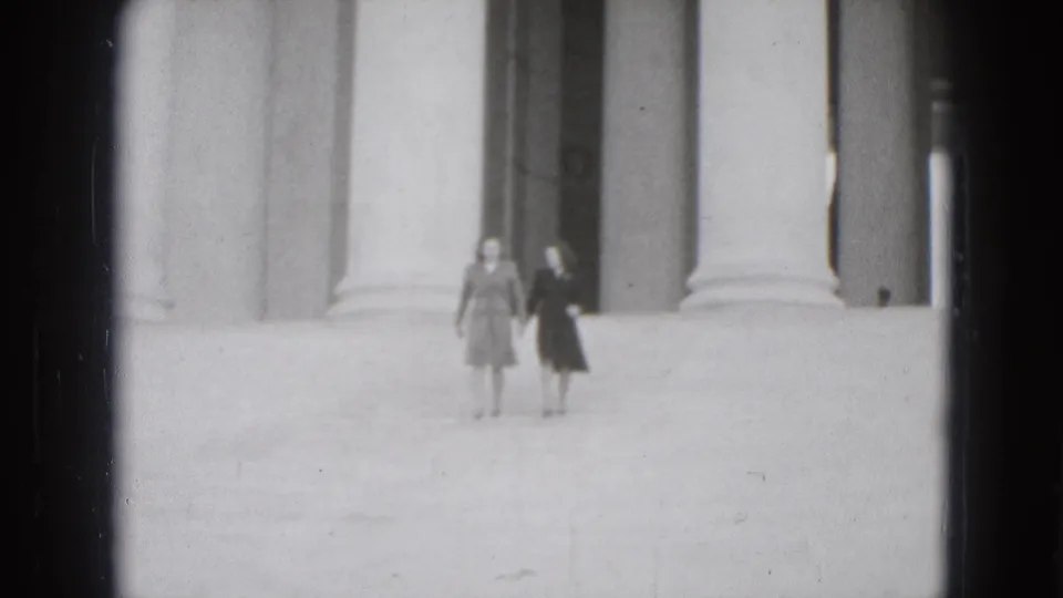Two women approach the camera on wide steps, with tall columns and dark recessed openings behind them in black-and-white 8mm footage.