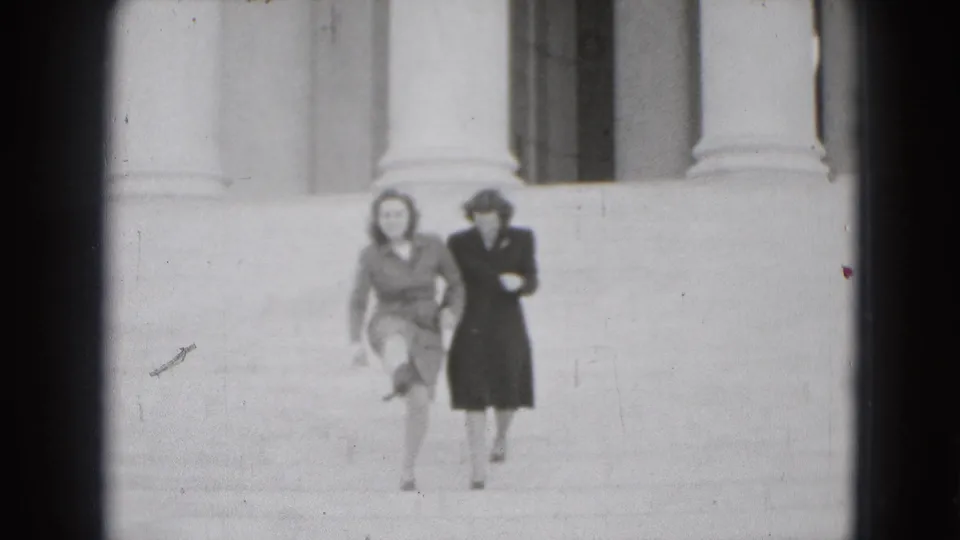 The two women are closer to camera while descending the stone steps, with one in a light suit and the other in a dark dress.