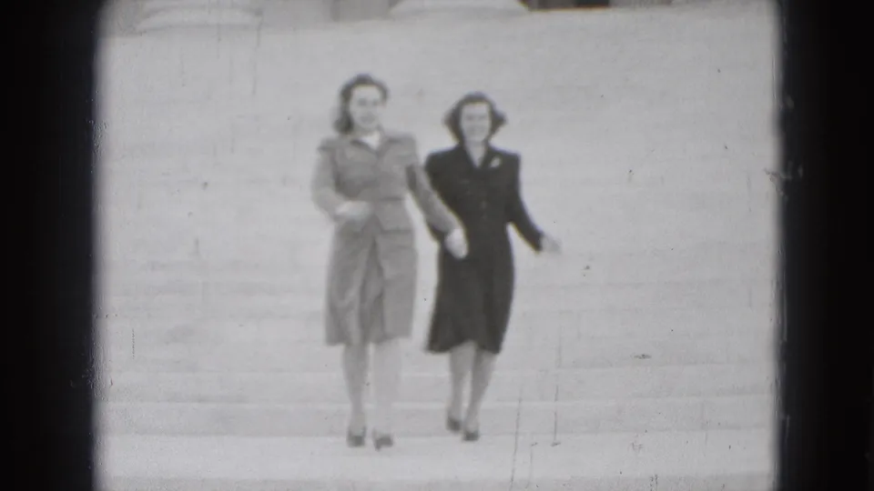 Two adult women in 1970s-style coats and heels walk side by side down wide stone steps in Washington, DC.