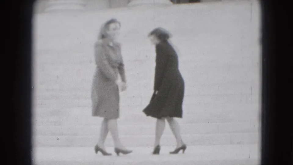 Two adult women in coats appear to turn toward each other on broad stone steps, with wind and motion softening the frame.