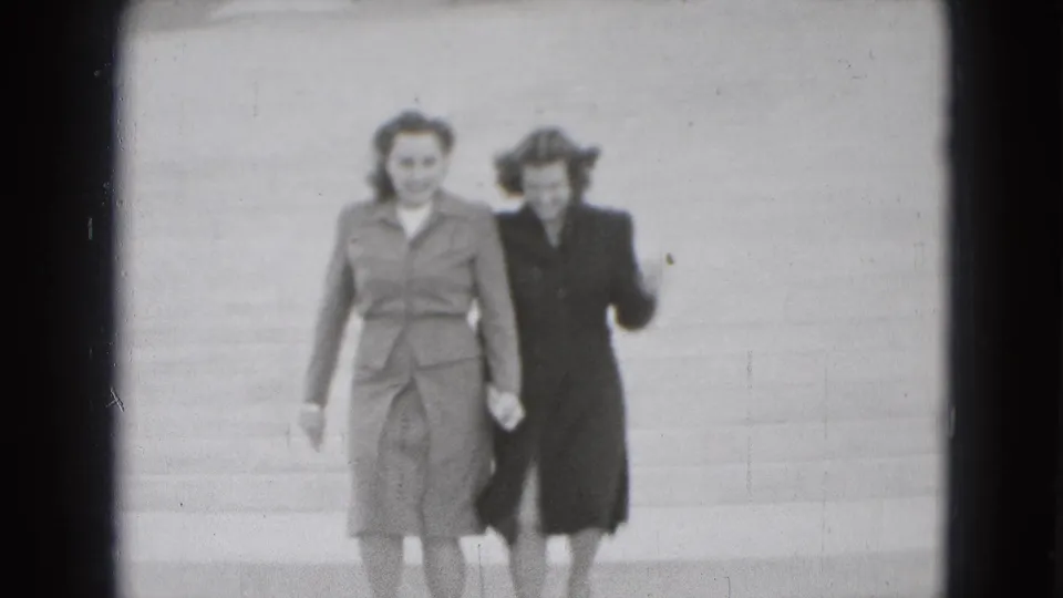 Two women in coats and heels hurry down wide outdoor steps, their hair and clothing showing wind and movement.