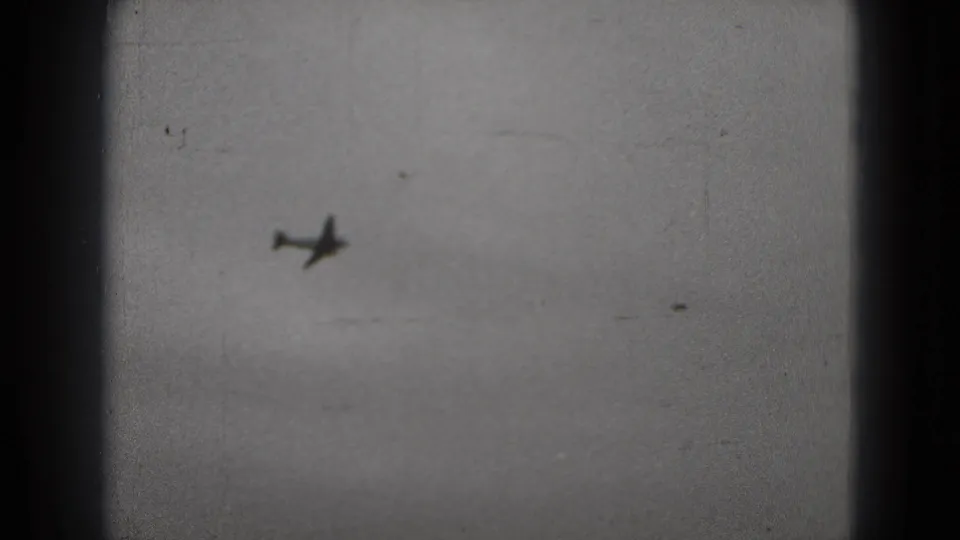1975 8mm frame showing a small airplane silhouette against cloudy sky, with scratches and dark viewfinder borders visible.