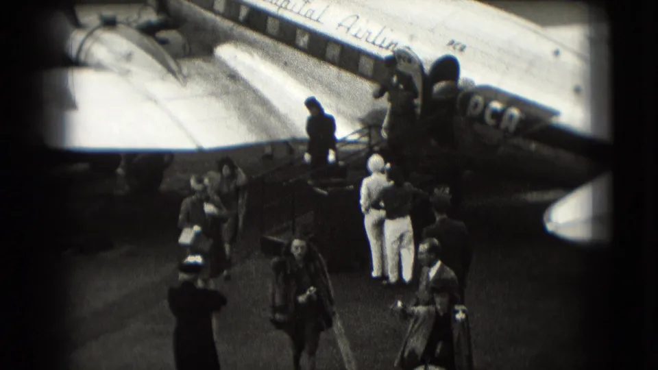 Black-and-white 8mm frame of 1975 Washington DC passengers and staff beside aircraft stairs on an airport tarmac.