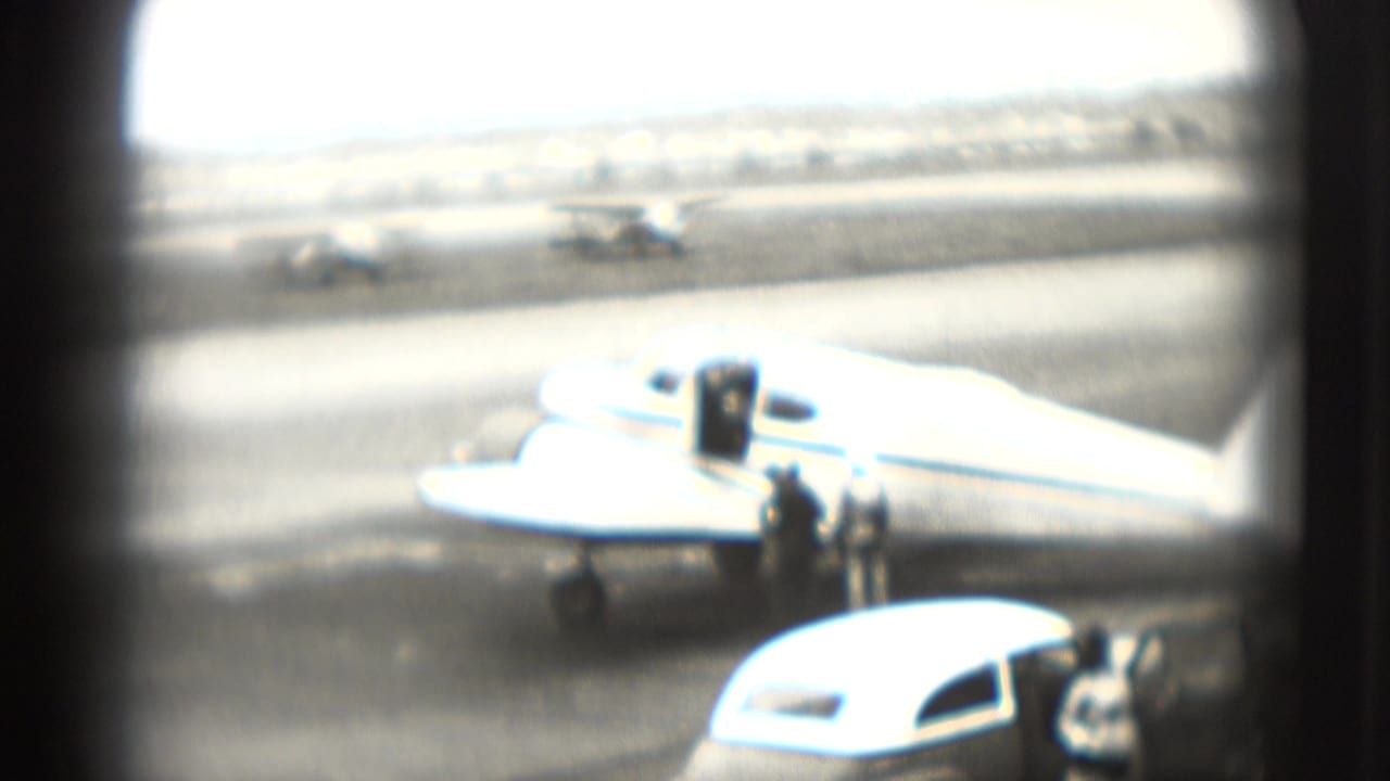 A small propeller plane is parked on a Washington, DC airport tarmac in 1975, with people and cars visible nearby.