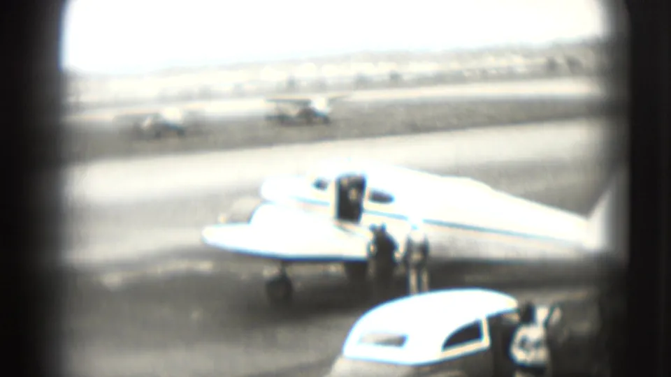 A small propeller plane is parked on a Washington, DC airport tarmac in 1975, with people and cars visible nearby.