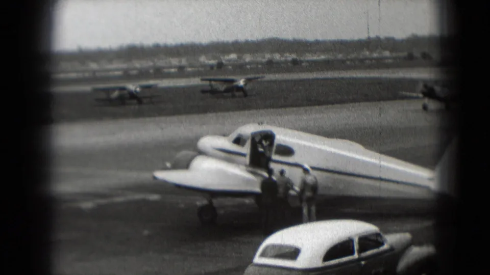 A small propeller plane remains parked on a Washington, DC tarmac in 1975, with people, cars, and other aircraft visible.