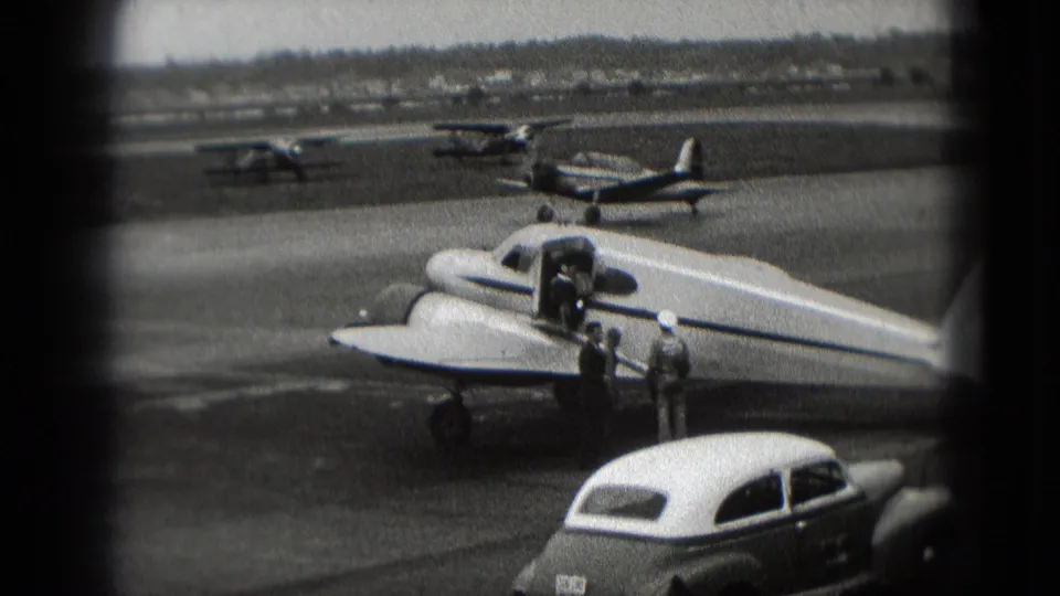 1975 Washington, DC airport home-movie frame showing a small propeller plane, open door, two adults, parked cars, and distant aircraft.