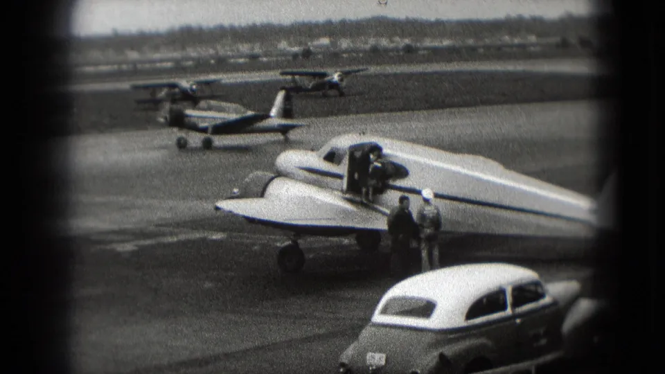 1975 Washington, DC tarmac view with a small propeller plane, adults near the wing, classic cars, and light aircraft in the background.