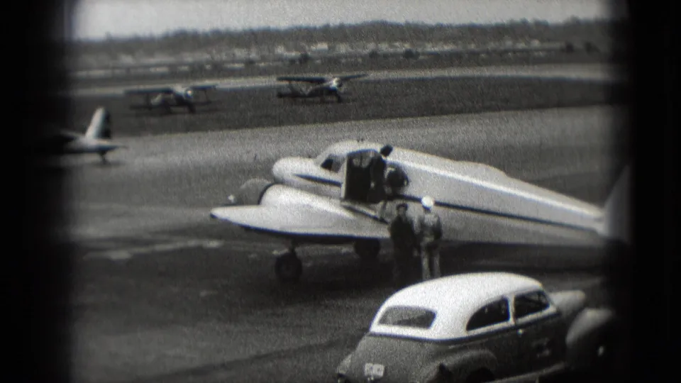 Washington, DC 1975 airport frame showing the nose and wing of a small plane, two adults, a foreground car, and distant aircraft.