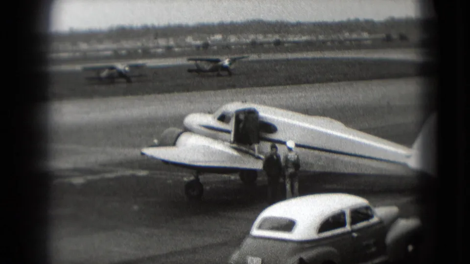 1975 Washington, DC home-movie still of a small plane on the tarmac, adults nearby, classic cars, and other aircraft beyond.